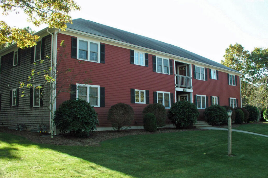 exterior view of a red two-story clapboard apartment building with a lush green lawn in front