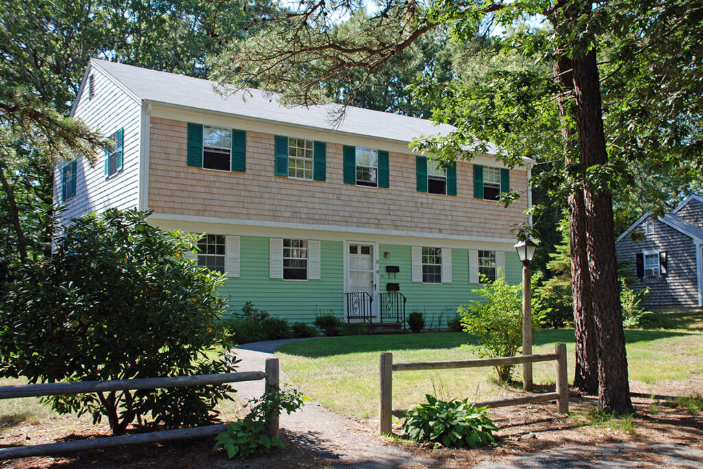 shingled Colonial-style house set back among trees on a sunny day